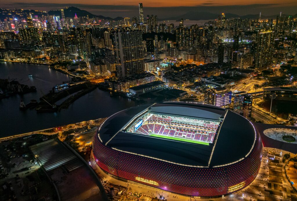 2025 MAMA Awards lineup Hong Kong — wide drone shot of Kai Tak Stadium with stage lighting tests at night.